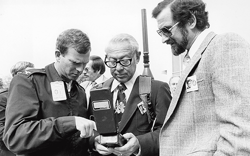 6)	B. P. (Pete) Leonard wears a Navstar backpack in this 1978 photo. Leonard, then vice president of Aerospace’s Navstar program group, is flanked by Col. Don Henderson (left) of the SAMSO Navstar program office and Ed Lassiter (right), principal director of Aerospace’s Satellite Navigation Systems Directorate.